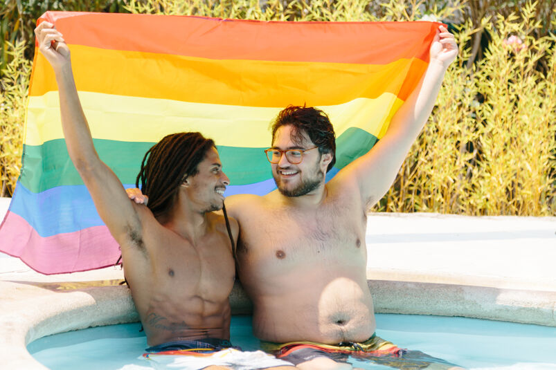 Two men in a hot tub smiling at each other and holding the rainbow flag.