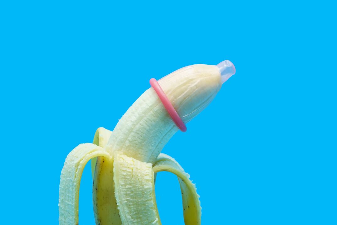 A peeled banana against a blue background, with a pink rimmed condom applied to its tip.