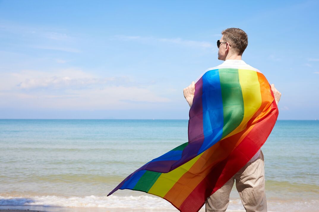 A man holding a Pride flag on the beach.