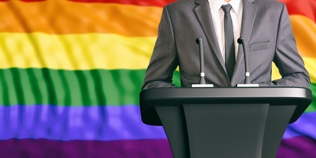 A suit at a podium in front of the Pride flag. 