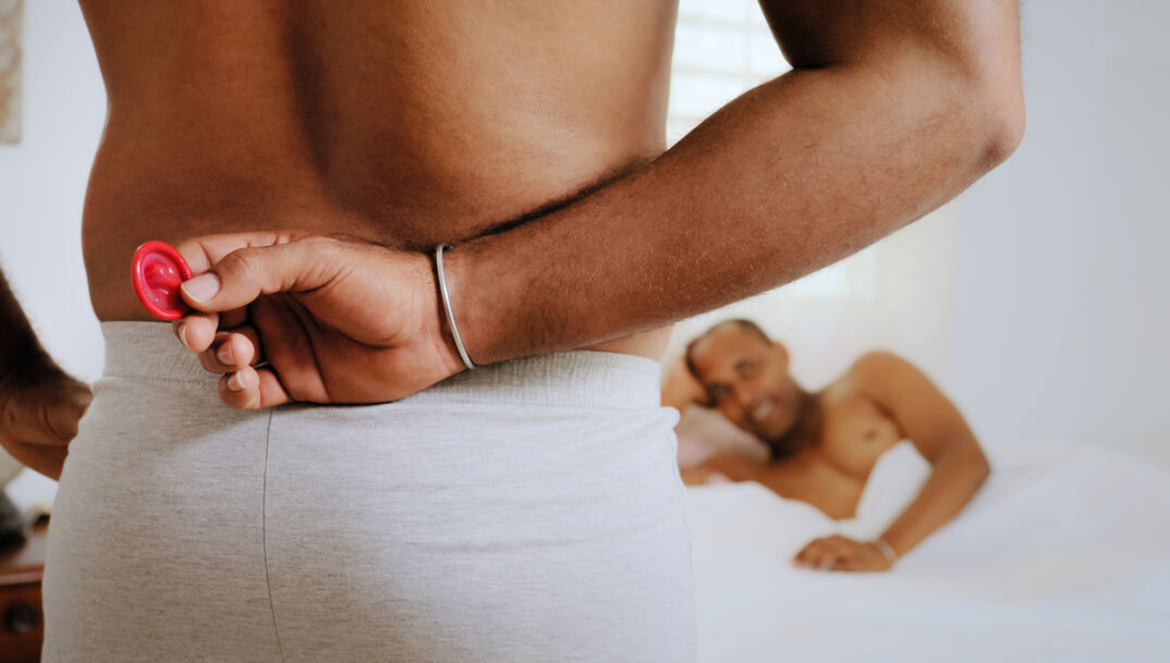 Back of man holding condom while another man lays in bed. 