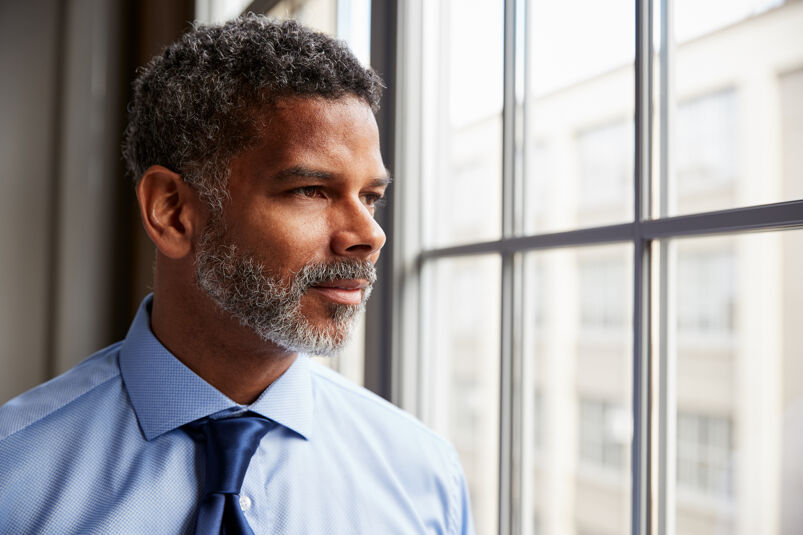 A black man with grey hair and beard is looking out a window. 
