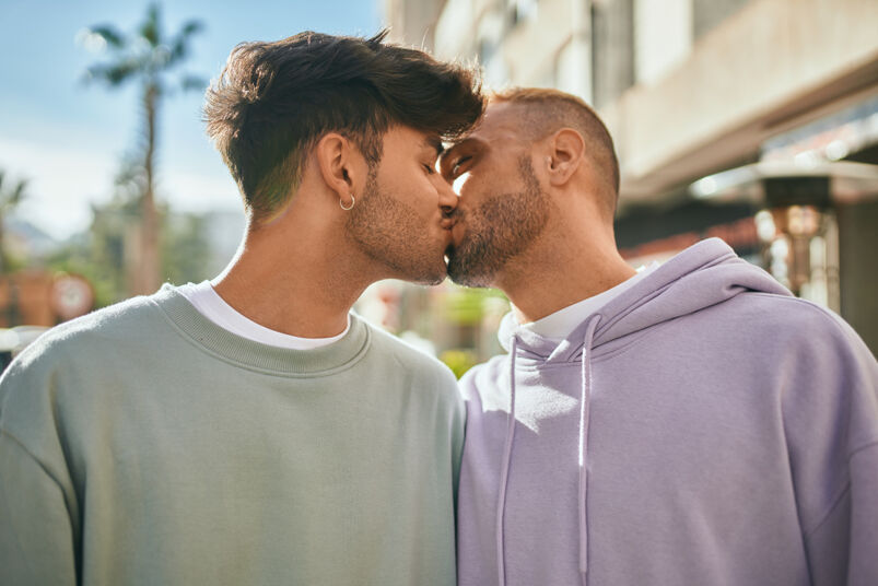 Straight guy dared to kiss gay friend prefers making out instead: A young, gay couple kissing in the city. One is wearing a light green hooded sweater, the other is wearing a matching hooded sweater, but in light purple. 