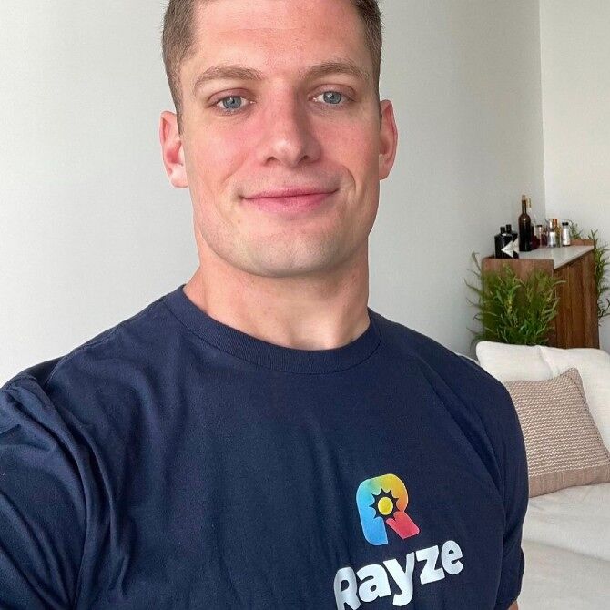 Carl Nassib standing in a living room in a black t-shirt with the lettering "Rayze" on his front. 