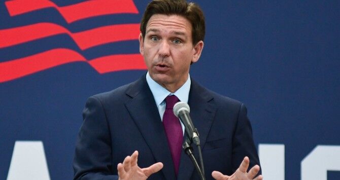 Ron DeSantis standing behind a podium wearing a dark suit jacket, blue shirt and purple tie. 