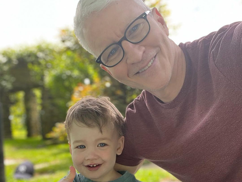 Anderson Cooper smiles taking a selfie with his son Wyatt, who is smiling holding a thumbs-up. Cooper wears black glasses and a maroon t-shirt. They pose in front of a lush green forest.