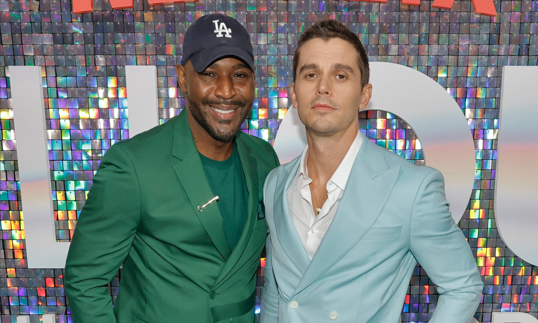 Karamo Brown, wearing a navy LA baseball hat, and a green blazer over a green tee, smiles with his arm around Antoni Porowski. Porowski wears a powder blue suit jacket over a white button down, open to reveal a gold necklace. He smirks with a short haircut.