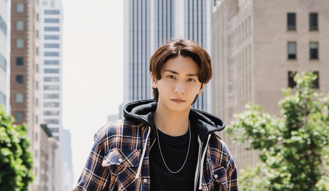 Shinjiro Atae, wearing a plaid hoodie unzipped over a black shirt, poses in the middle of a city street. His hair is parted in the middle and he smiles softly.