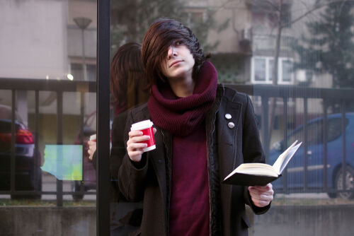 Emo boy with book and coffee is standing outdoors at the street and resting.