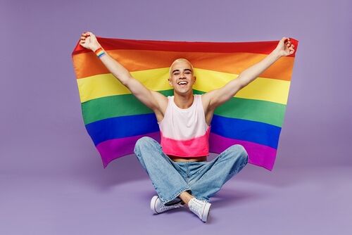 Twink: Full body happy young latin gay man 20s with make up wearing bright pink top sitting looking camera isolated on plain pastel purple background studio portrait. 