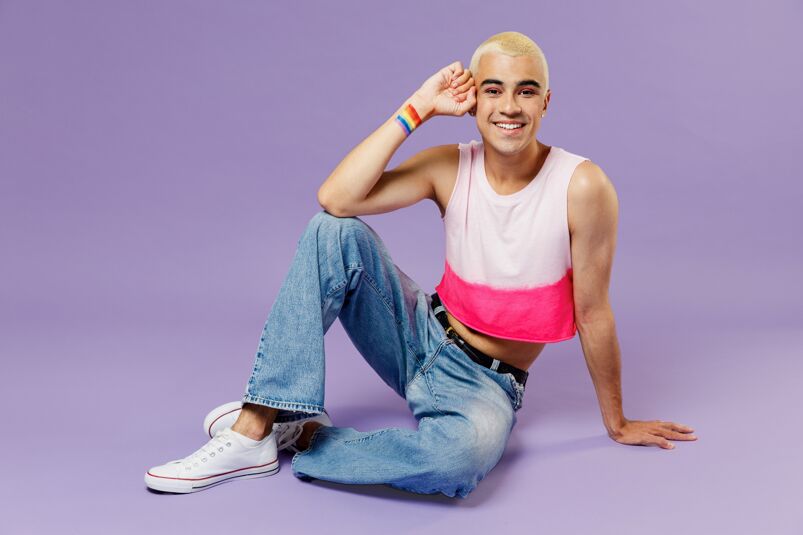 Straight equivalent to gay terms: Full body happy young latin gay man 20s with make up wearing bright pink top sitting looking camera isolated on plain pastel purple background studio portrait. 