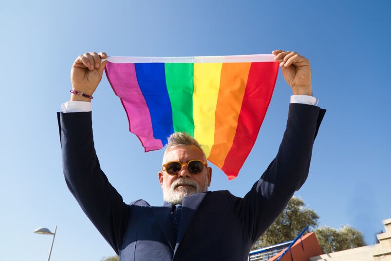 An older man wearing sunglasses and a suit waving a Rainbow flag. 