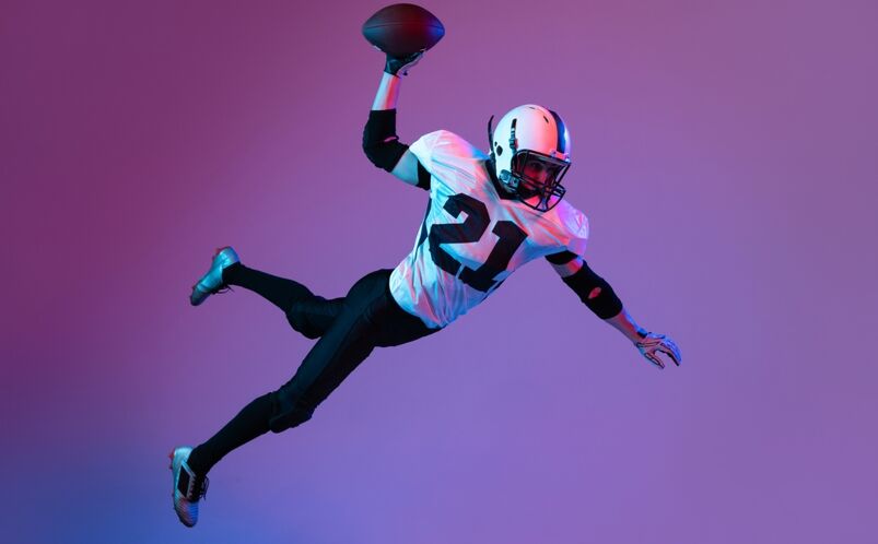 Portrait of american football player in motion, catching ball in a jump iisolated over purple background in neon light
