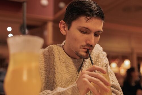 Portrait of one young handsome brunette caucasian man in knitted sweater drinking a cocktail with a straw from a glass, sitting at a table in a restaurant, close-up side view. 