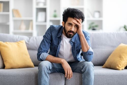 Portrait Of Depressed Young Indian Man Sitting On Couch At Home