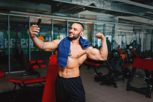 Straight equivalent to gay terms: A gorgeous and muscular man flexing and showing off his arm muscles on a blurred gym background. A manly bodybuilder with blue towel, headphones and smartphone taking selfies of his hot, fit body.