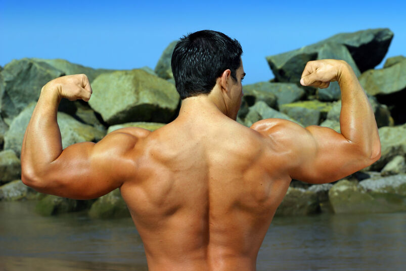 Spornosexual body builder flexing his back by the ocean with rocks in the background.