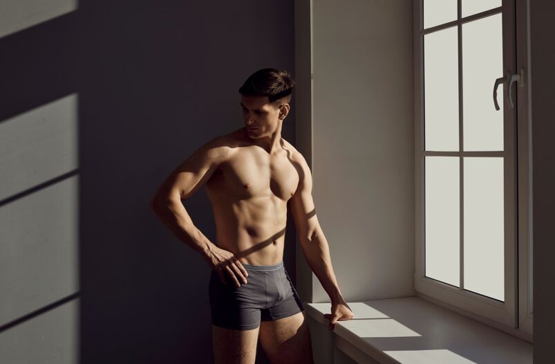 Sexy young Caucasian man in underwear stand near window at home. Muscular toned millennial male model in briefs indoors. Fit athletic handsome model show naked torso. Fashion and clothing.
