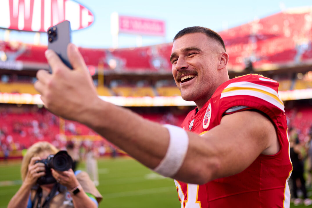 Travis Kelce wearing his red Chiefs uniform on the field and taking a selfie. 