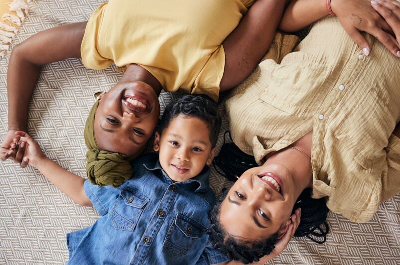 Gender neutral gay baby names: LGBT, happy portrait and relax family, child or people care, smile and lesbian bond together, lying and on floor carpet. Mothers face, top view kid and non binary parents, homosexual mom or gay women