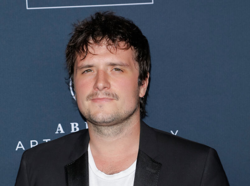 Josh Hutcherson, with dark messy hair, a mustache, and five o' clock shadow, softly smiles in front of a step-and-repeat. He wears a black blazer over a white tee.