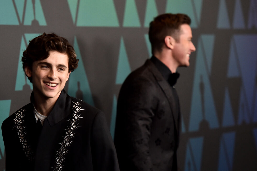 Timothee Chalamet, wearing a black blazer over a white t-shirt smiles in front of a step and repeat at the camera in the foreground. Behind him in the background, Armie Hammer, also dressed in black, smiles while facing the opposite direction. He is slightly blurred.