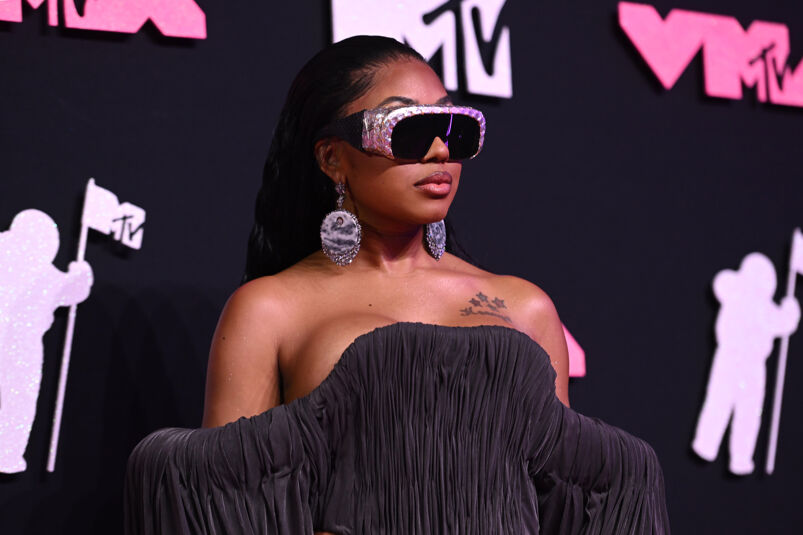 Rapper Yung Miami stands in front of a black and pink step and repeat the MTV VMAs. She wears sunglasses that wrap around her faces with pink trim, low-hanging pink leopard print earrings, and a shaped strapless black gown that starts right at her cleavage. 