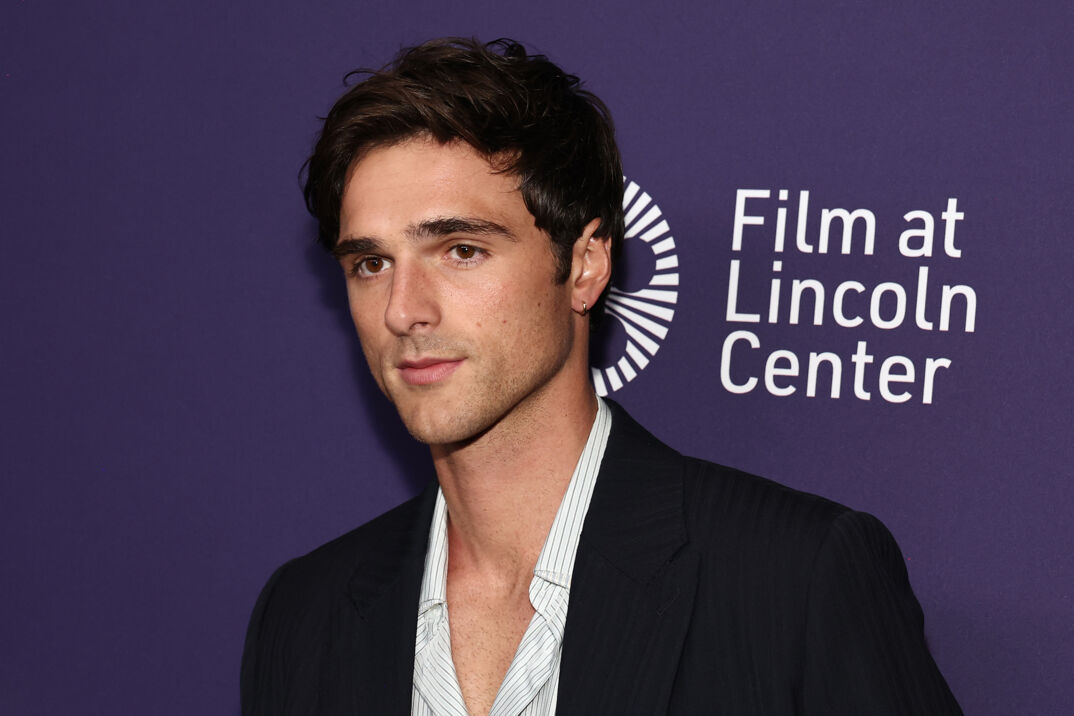 Jacob Elordi, wearing a striped t-shirt unbuttoned to reveal his chest hair and a black blazer looks away from the camera in front of a step and repeat. The background reads "Film at Lincoln Center."
