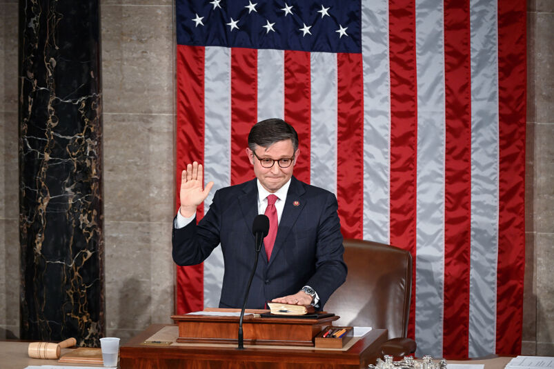 Speaker Mike Johnson taking the oath of office behind the House podium, standing in front of a giant American flag. 