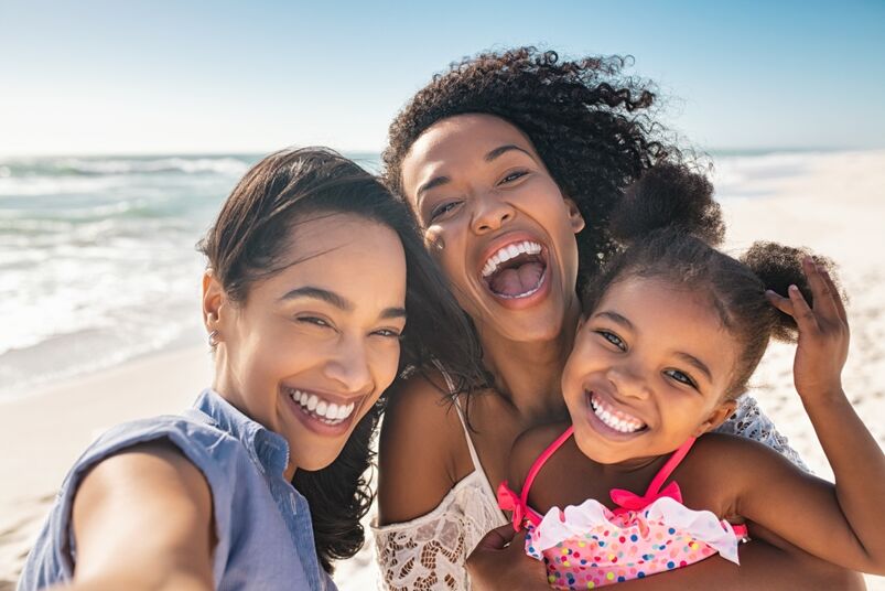 Girl gay baby names: Portrait of smiling young african american woman with child taking selfie at beach with her best friend. Cheerful multiethnic gay couple enjoying at beach with daughter during summer holiday.