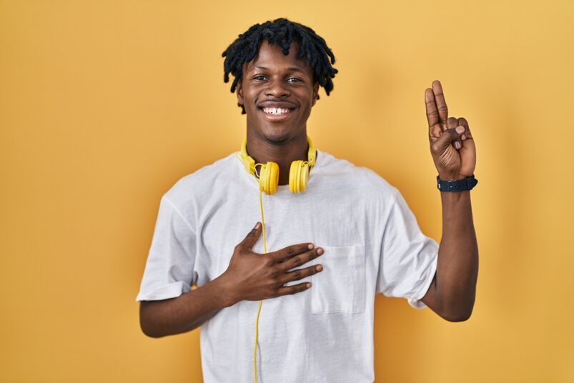 Young Black man with dreadlocks standing over yellow background smiling swearing with hand on chest and fingers up, making a loyalty promise oath.