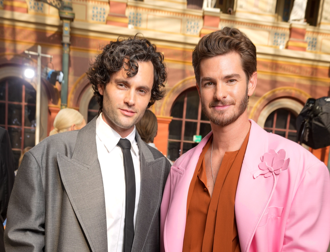 Penn Badgley, wearing a gray overcoat on top of a white button down shirt with a dark tie, stands next to Andrew Garfield. Garfield wears an orange, untied pussy bow blouse and a pink overcoat. They stand smiling in front of a crowd at the Valentino 2023 fashion show.