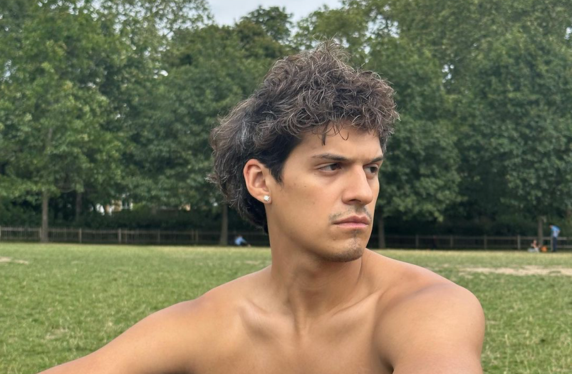 Omar Apollo sits shirtless with messy hair in a lush green park. His arms are on his knees and he looks off pensively.