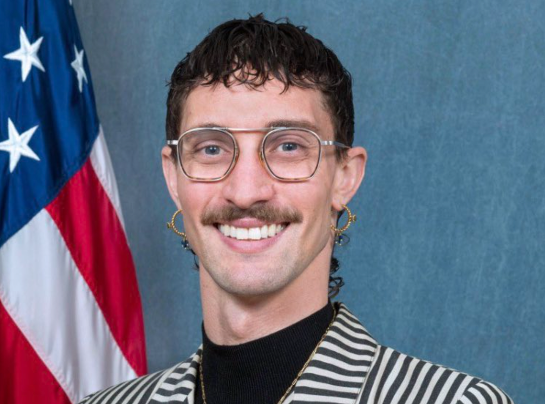 Tyler Cherry wears wire-rimmed glasses, golden hoop earrings and a golden necklace over a black turtleneck and black-and-white striped suit jacket. They have a shaggy brown haircut and smile in front of a blue background and United States flag in their DC headshot.