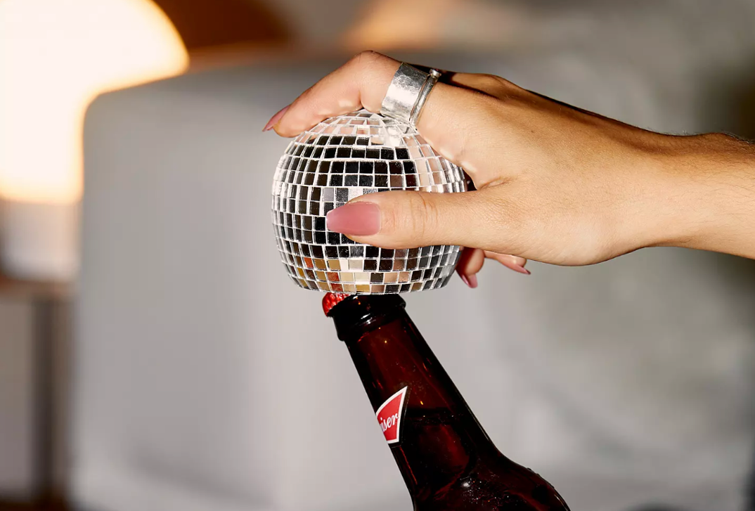 A hand with pink nails sticks its pointer finger in a handsize disco ball with the bottom cutoff that allows it to open beer bottles. A Budweiser beer bottle is positioned underneath it.