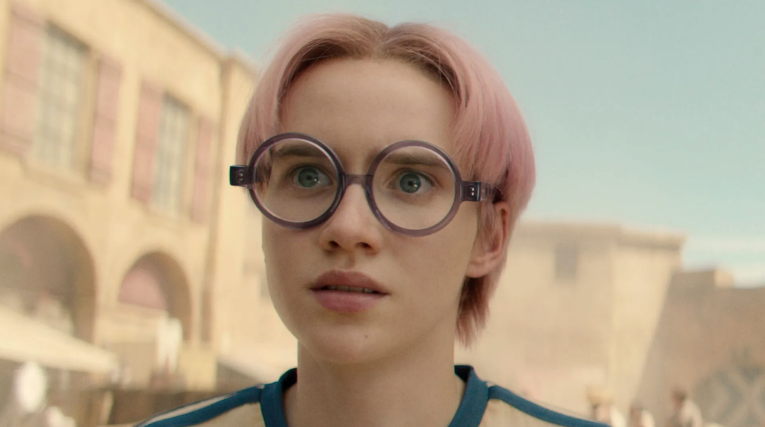 Morgan Davies, wearing circular glasses, stares incredulously off camera in a scene from Netflix's 'One Piece.' He has light pink hair parted in the middle and wears a blue and white t-shirt.