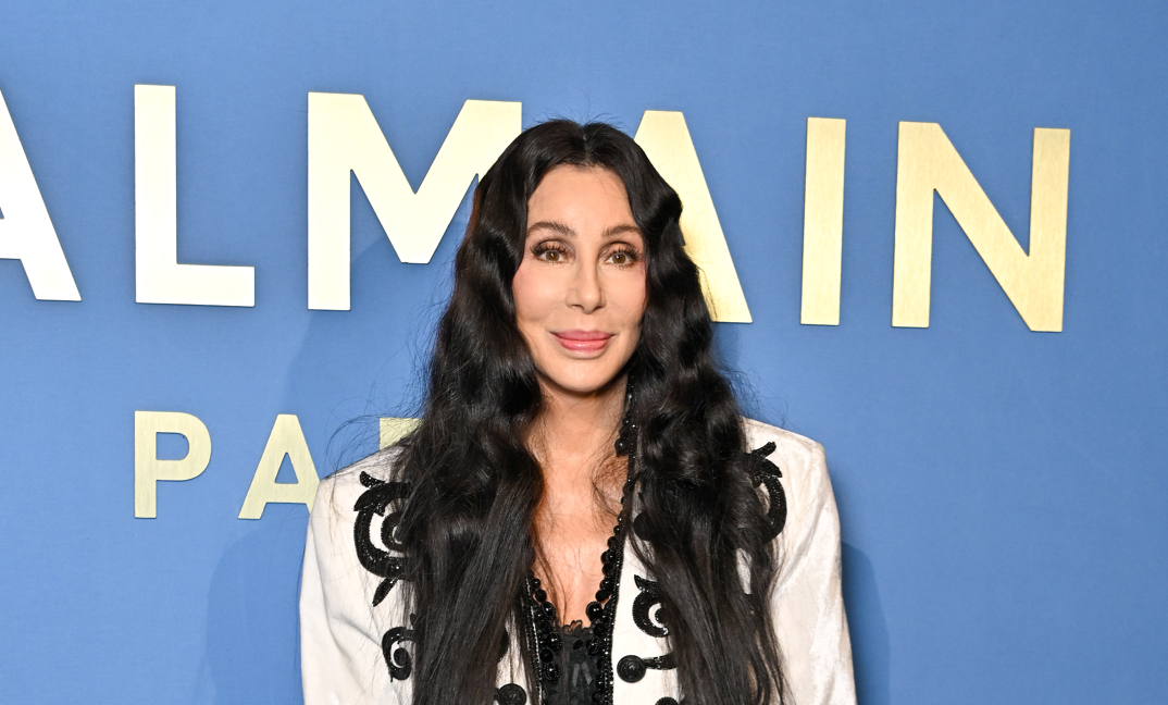 Cher, sporting long black hair and pink lipstick, stands smiling in front of a blue background at the Almain Paris Fashion Week step-and-repeat. She wears a white blazer adorned with black sparkling swirls, opened to reveal a black tank-top.