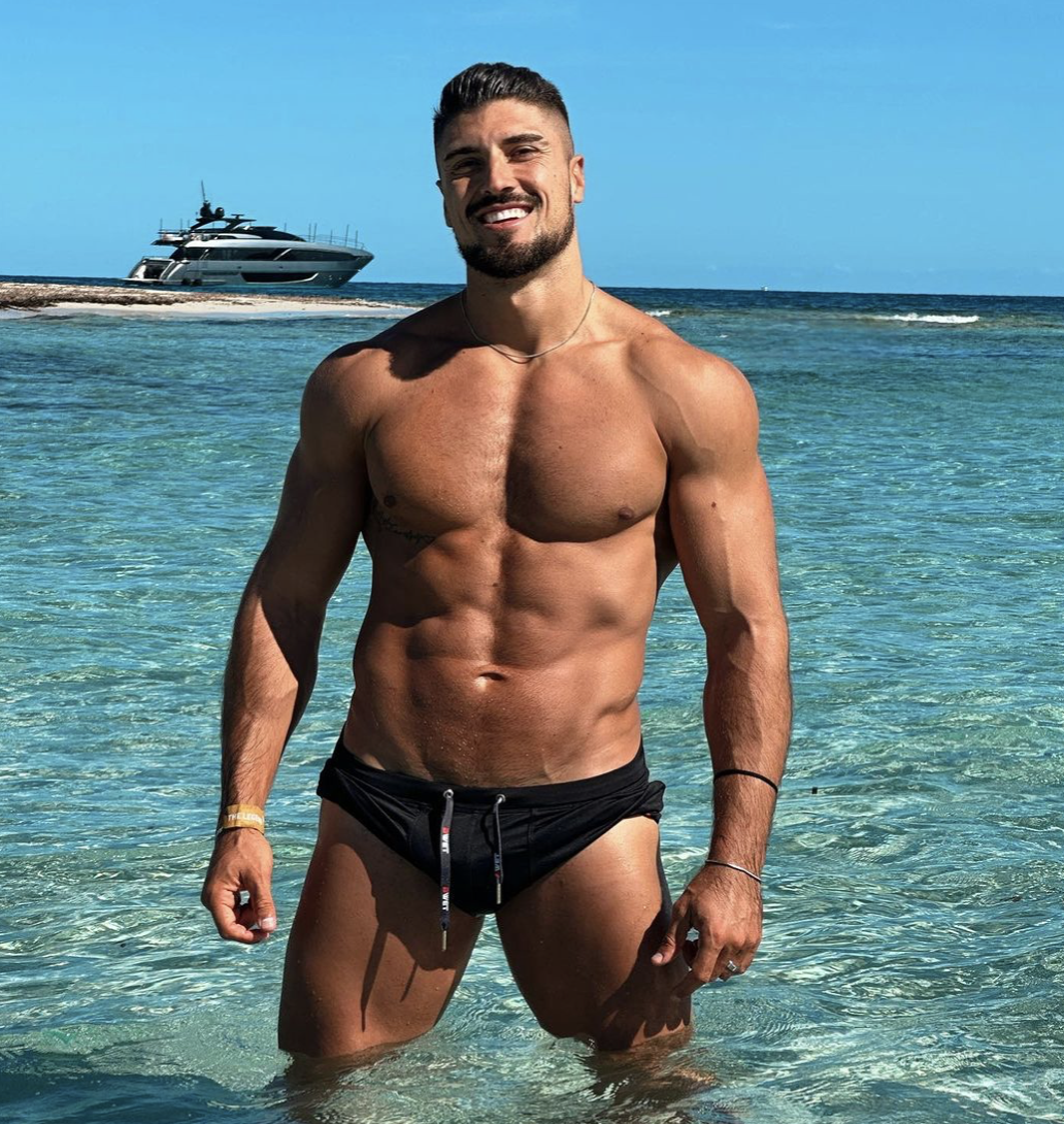 Miguel Gonzalez standing shirtless in the ocean in a black speedo. 