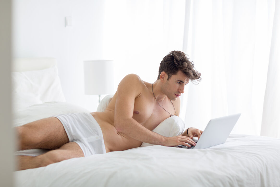 A shirtless man in white briefs lays on a white bed in front of white curtains and a white lamp. He wears a necklaces and leans over a pillow while he works on a gray laptop intently.