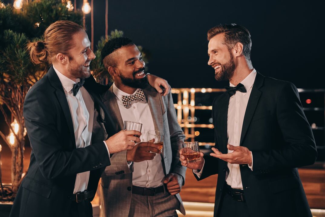 Three men in suits laughing and toasting with bourbon drinks.