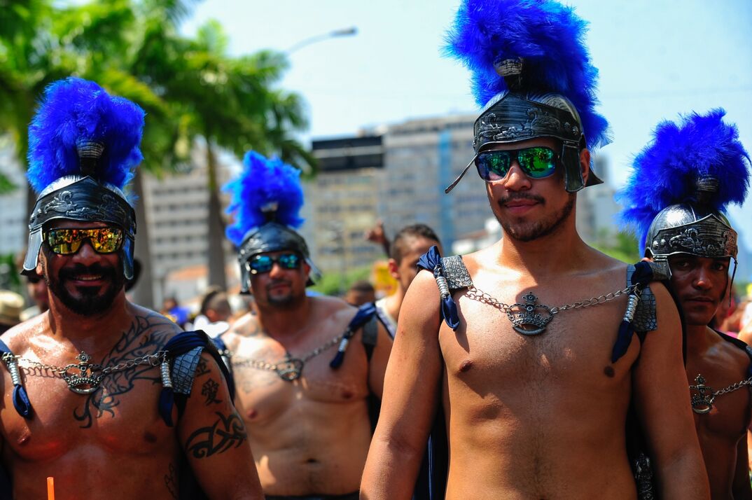 Muscular, shirtless men walk around a downtown area during the daytime. They have Roman gladiator helmets on featuring flamboyant blue feathers and wear sunglasses.