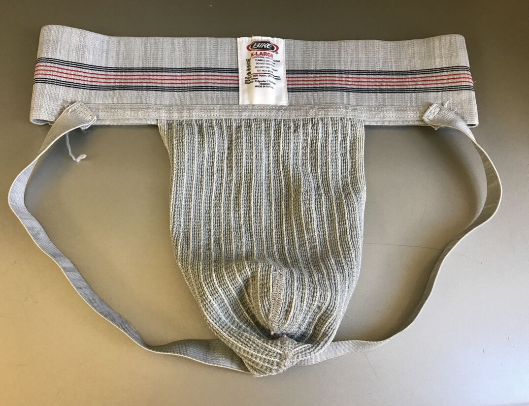 A gray jockstrap lays on a off white table. A tag on the jockstrap reads "X-Large" and the brand "BIKE." There's a red and blue line across the waist.