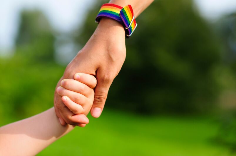 The Queerty guide to gay baby names: Parent holds the hand of a small child. mother holding baby's hand. rainbow LGBTQ+ bracelet on parents hand.