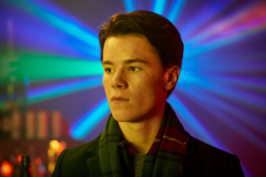 'Young Royals' star Edvin Ryding wears a scarg and looks to the side as colorful lights shine behind him.