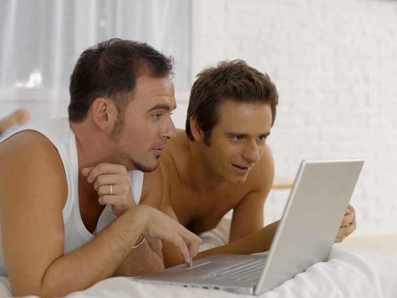 Two men sit on a white bed looking at a gray laptop. The man on the right wears a wedding ring, white tank top, and has dark hair and a goatee. Immediately to his right is a shirtless man with spiky brunette hair smiling at the screen.