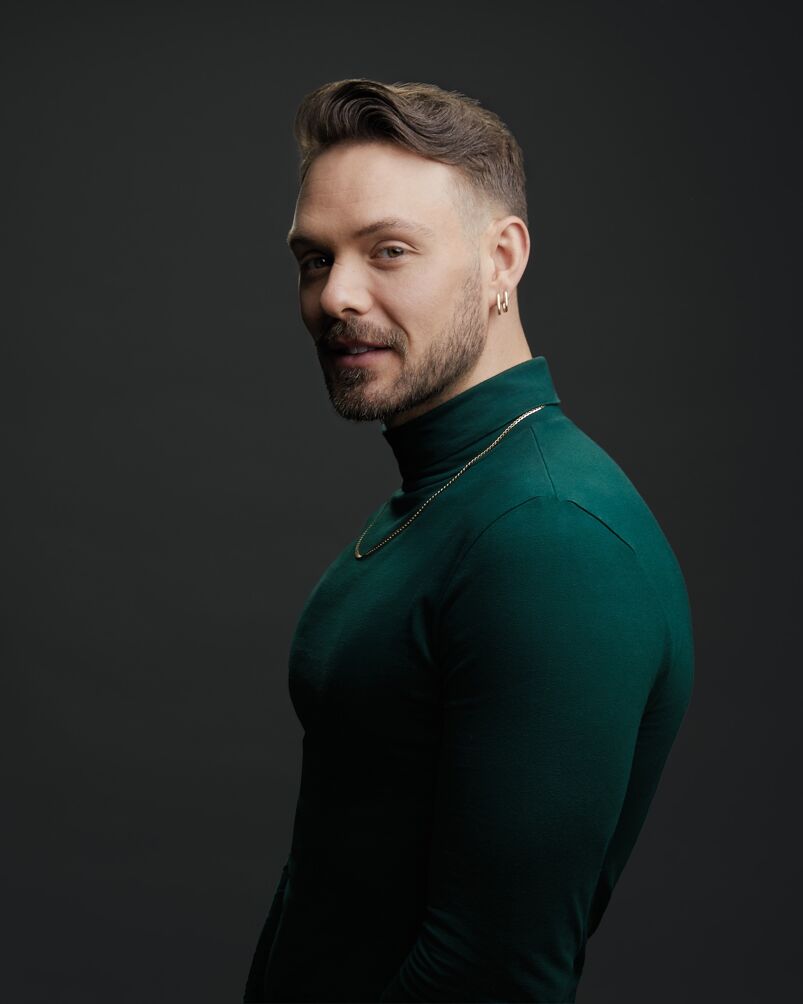 John Whaite poses in a green turtleneck and gold chain in front of a grey backdrop.