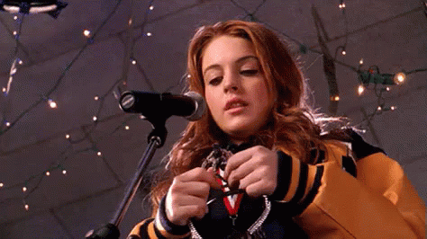 Lindsay Lohan, wearing a blue and yellow varsity jacket, breaks a plastic crown in half behind a microphone in a scene from 'Mean Girls.'