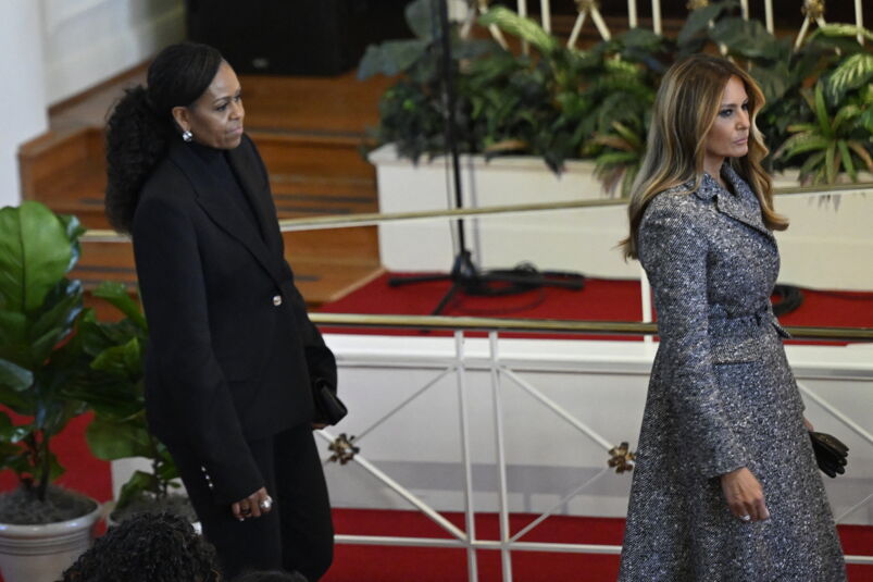 Michelle Obama, wearing black, standing behind Melania Trump in a gray overcoat. 