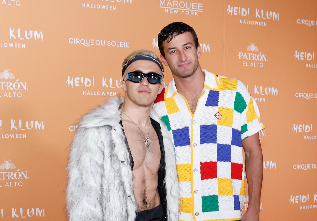 Chris Olsen, wearing a furry coat open to reveal a ripped chest, a necklace, black sunglasses, and a bandana wrapped around his bleached hair stands next to Patrick Johnson. Johnson smiles softly in a red, blue, yellow, and green checkerboard white t-shirt like Allan from 'Barbie.' They stand in front of an orange step and repeat for Heidi Klum's Halloween party.