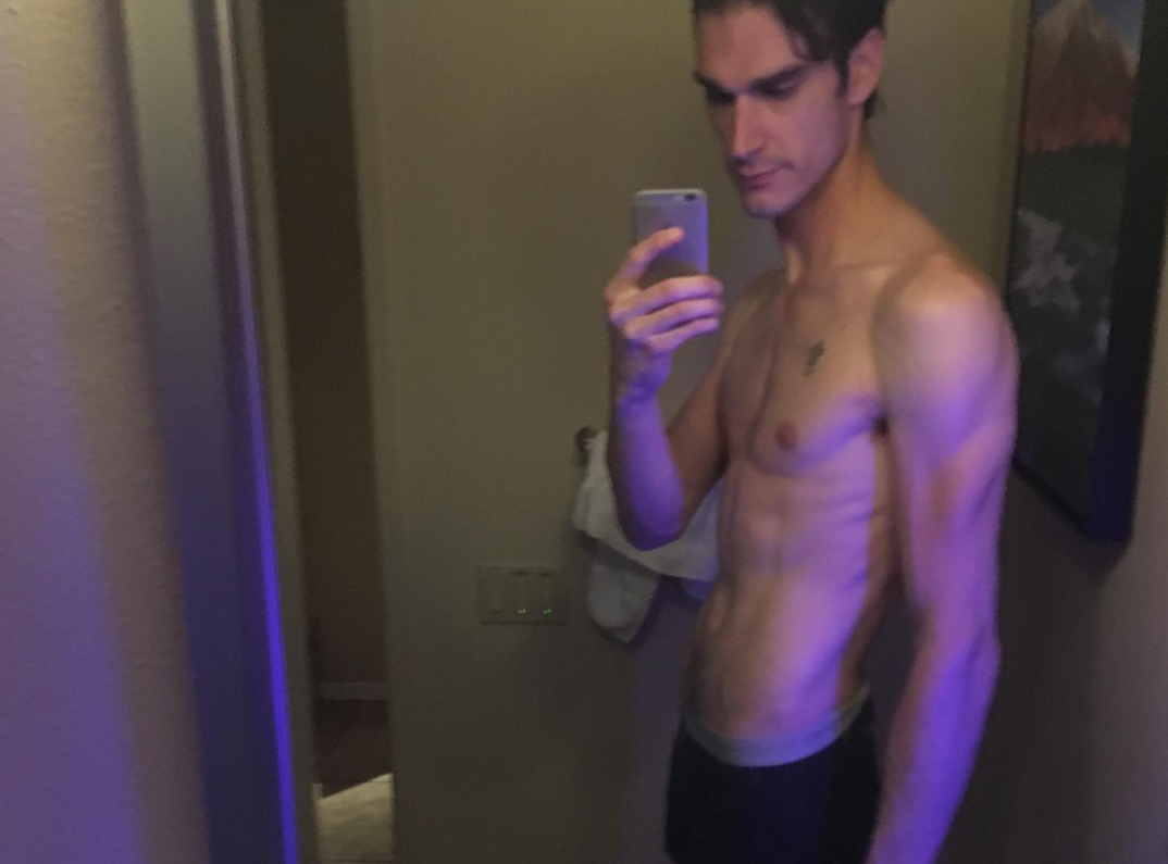 Marston Hefner, who is tall and skinny with dark brown hair parted in the middle, stands shirtless in front of a bathroom mirror. He holds a gray iPhone up to capture the photo. A gray boxer waistband peeks out of his black pants.